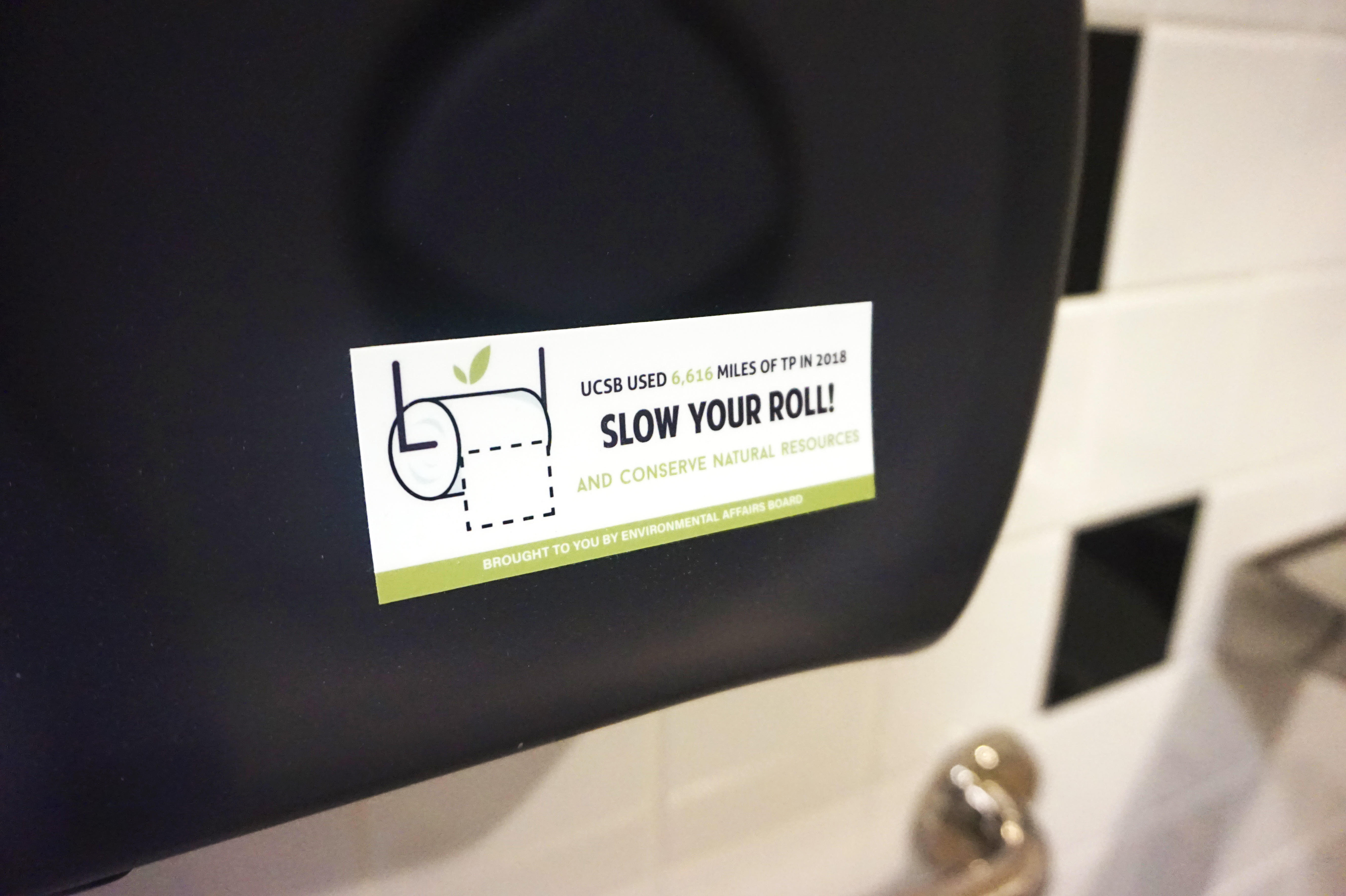 A photograph of a sticker on top of a toilet paper dispenser that reads 'Slow Your Roll!